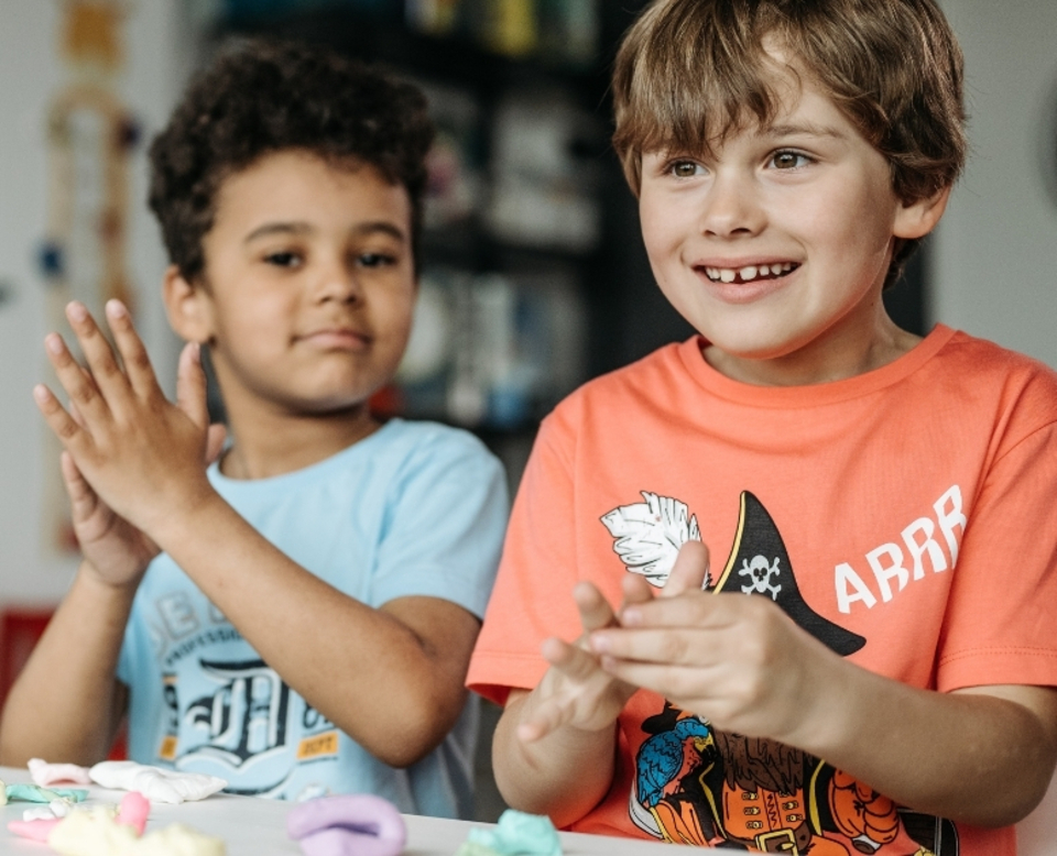 Two young boys smiling and playing with colorful modeling clay at a table. One wears a light blue shirt, the other an orange shirt with a pirate graphic.